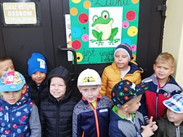 Dzieci w ubraniach wierzchnich pozują na tle zielonego plakatu z żabką 
