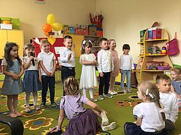 8 dzieci stoi na środku sali przedszkolnej, dziewczynka trzyma mikrofon w ręce