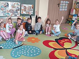 Dzieci siedzą na dywanie i podnoszą ręce w górę 