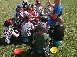Dzieci siedzą na trawie i słuchają trenera