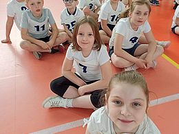 dzieci siedzą po turecku na hali sportowej i patrzą w obiektyw
